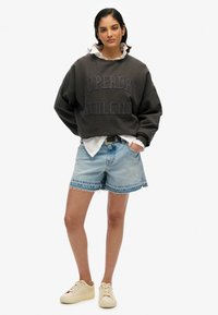 Sweatshirt gris avec le texte "SUPERDRY ATHLETIC", associé à un short en denim bleu clair avec un ourlet effiloché, et des baskets beiges.