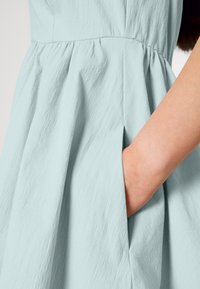 Ljust blå klänning i texturerad tyg, med en samlad midja och sidofickor synliga till höger.