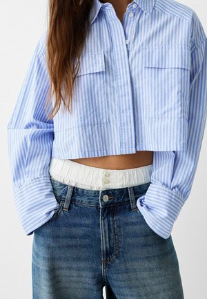 Frau trägt ein hellblau-weiß gestreiftes, bauchfreies Hemd mit langen Ärmeln, das in hoch taillierte blaue Jeans gesteckt ist.