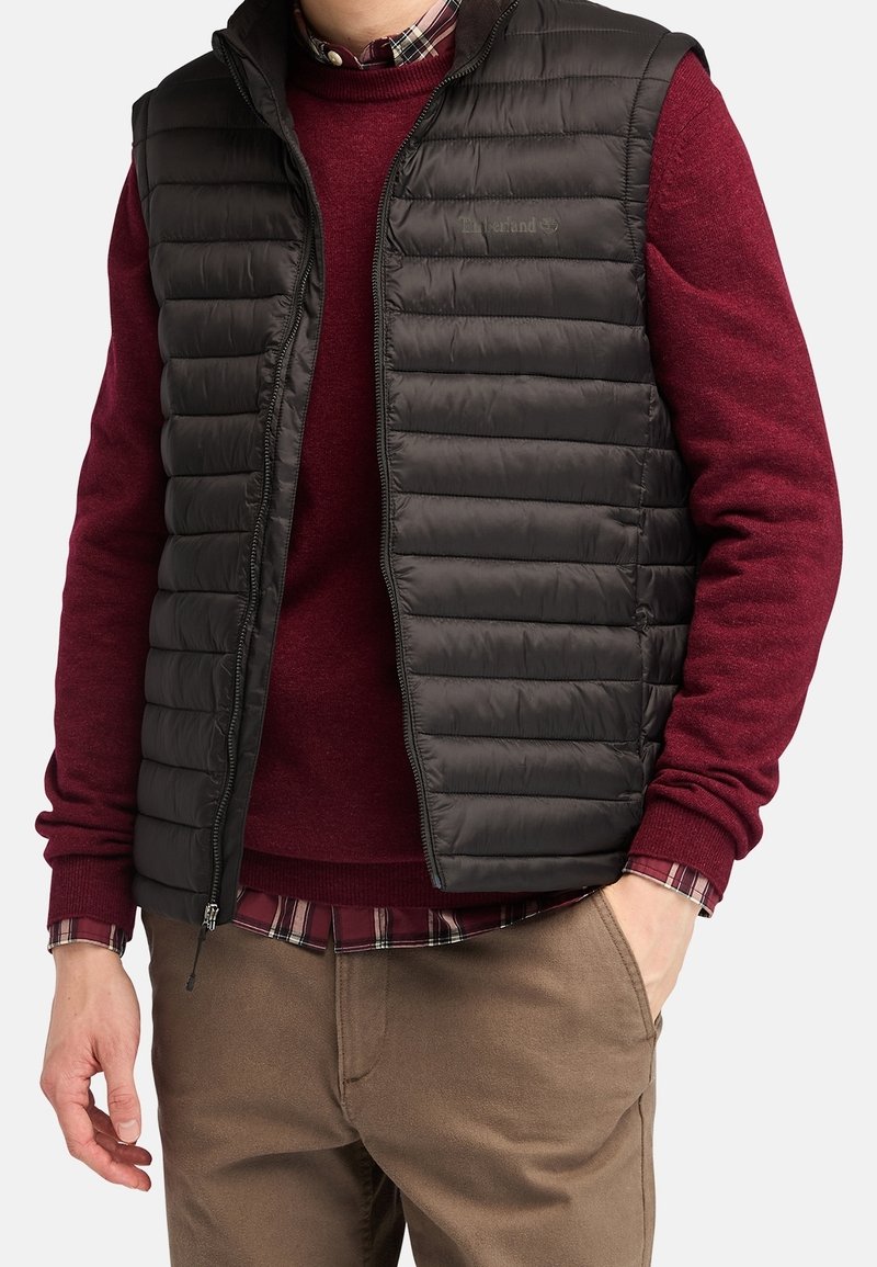 Homme portant une veste sans manches matelassée noire sur un pull bordeaux et une chemise à carreaux, avec un pantalon marron, main dans la poche, devant un fond uni.