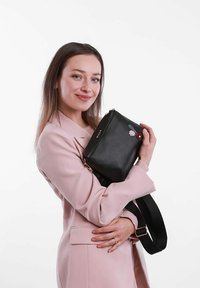 Černá kožená kabelka s květinovou výšivkou, obdélníkového tvaru, se zlatými doplňky a nastavitelným ramenním popruhem. Modelka pózuje s kabelkou.