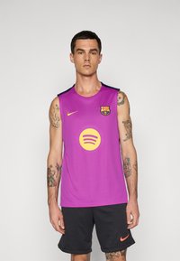 Egy férfi lila ujjatlan FC Barcelona sportmezben, sárga logókkal és fekete Nike sorttal áll egy egyszínű háttér előtt.