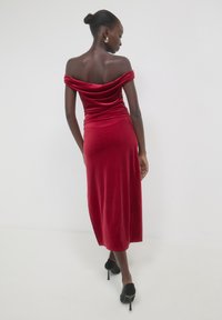 Rotes Samtkleid mit schulterfreiem Schnitt, drapiertem Ausschnitt, figurbetontem Silhouette und knielanger Länge. Getragen mit schwarzen High Heels.