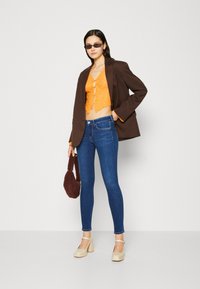 Blazer marrón, top corto de encaje naranja con botones, jeans skinny azul oscuro, zapatos de tacón crema y bolso texturizado burdeos.