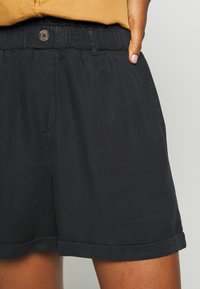 Pantalones cortos negros con cintura elástica, dobladillo cortado y un bolsillo delantero. Hechos de una tela ligera con una textura mate.