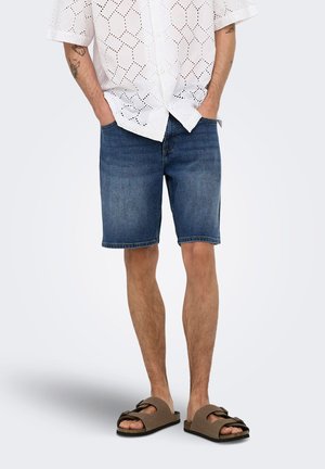 Uomo che indossa pantaloncini di jeans blu, camicia bianca a maniche corte con motivo e sandali marroni con due cinturini, in piedi con le mani nelle tasche.