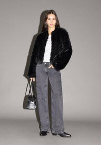 Schwarze Kunstfelljacke über einem weißen Hemd, kombiniert mit grauen hochgeschnittenen Jeans. Das Model hält eine kleine schwarze Tasche und trägt schwarze Schuhe.