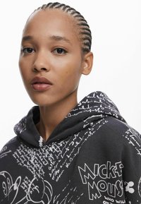 Zwarte hoodie met witte Mickey Mouse graphics en handgeschreven tekstpatronen. Het materiaal lijkt zacht en heeft een voorzak en een capuchon met trekkoord.