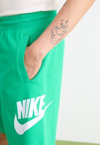 Shorts en coton vert avec une poche latérale, arborant un grand logo Nike blanc. Le tissu est lisse et présente un design décontracté et sportif.