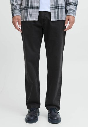 Man wearing black straight-leg pants, black shoes, white shirt, and gray plaid jacket standing against a plain background.