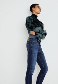 Mørkegrønn ribbestrikket genser med høy hals, kombinert med tettsittende mørkeblå jeans med en brun etikett på livlinen. Silversolbriller brukt.