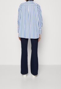 Camisa a rayas azul y blanca con un corte relajado, mangas largas y un detalle abotonado en la parte posterior, combinada con pantalones oscuros de campana y zapatos blancos.