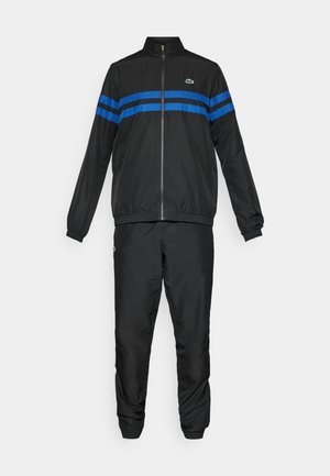 Training suit negru cu jachetă cu fermoar pe toată lungimea, având două dungi orizontale albastre și pantaloni asortați, cu manșete elastice și logo-uri Lacoste.