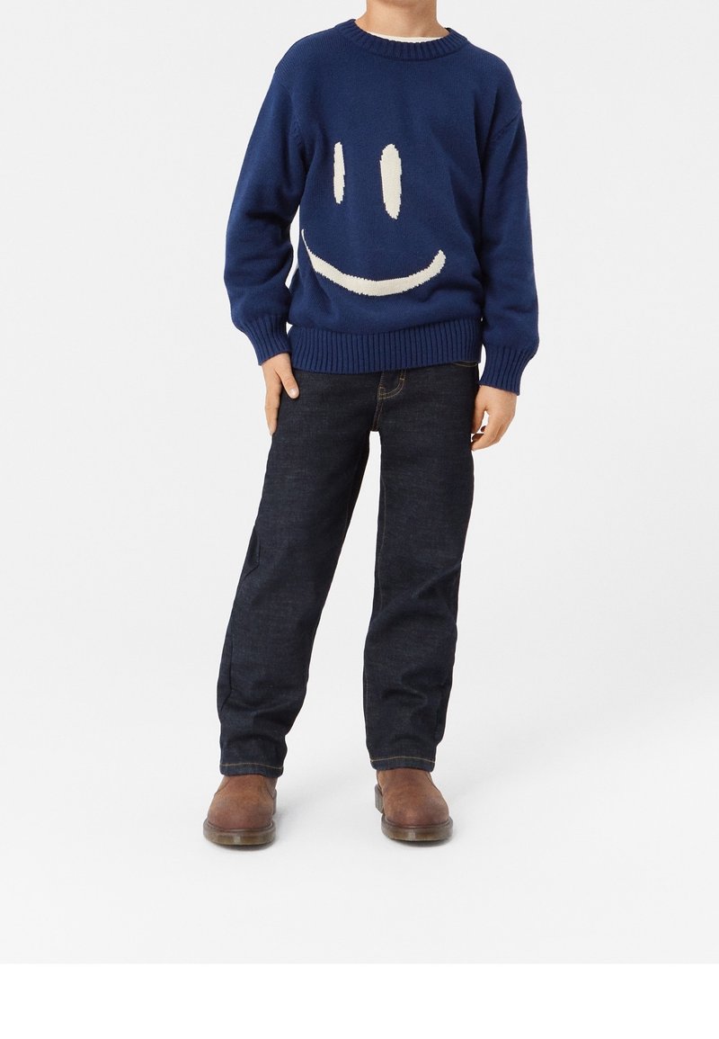 Blå stickad tröja med krämfärgad smiley-ansiktsdesign, ribbade ärmar, tillsammans med mörka denimjeans och bruna stövlar, stående mot en enkel bakgrund.