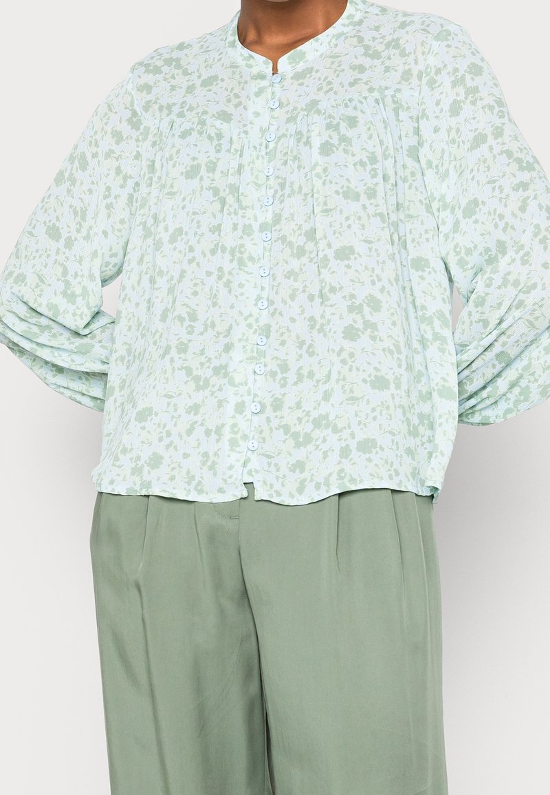 Chemisier à boutons à fleurs vert clair avec manches bouffantes longues, associé à un pantalon ample taille haute vert sauge.