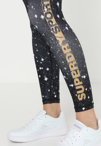 Czarne legginsy z gwiezdnym wzorem i złotym logo "SUPERDRY" po boku, w połączeniu z białymi butami sportowymi na platformie.