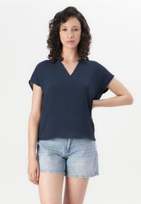 Navyblaues Kurzarmoberteil mit V-Ausschnitt und strukturiertem Stoff; kombiniert mit hellen Jeansshorts. Schlichte, lässige Passform und entspanntes Design.