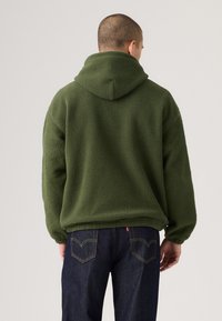 Green fleece hoodie with a textured surface, featuring a large hood and dropped shoulders, paired with dark denim jeans.