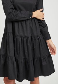 Schwarzes langärmliges Kleid mit einem gestuften Rüschendesign. Aus leichtem Stoff gefertigt, mit einer Knopfdetail am Handgelenk.