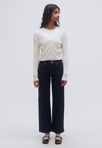 Ελαφρώς μπεζ μακρυμάνικη μπλούζα με κόμπο στο μπροστινό μέρος, συνδυασμένη με τζιν παντελόνι ψηλόμεσο, φαρδύ και σκουρομπλε, και καφέ παπούτσια. Απλό φόντο.