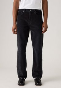 Zwarte corduroy broek met een rechte pasvorm, vijf zakken, metalen knoopsluiting en geribbelde textuur, gedragen met zwarte schoenen en een wit shirt.