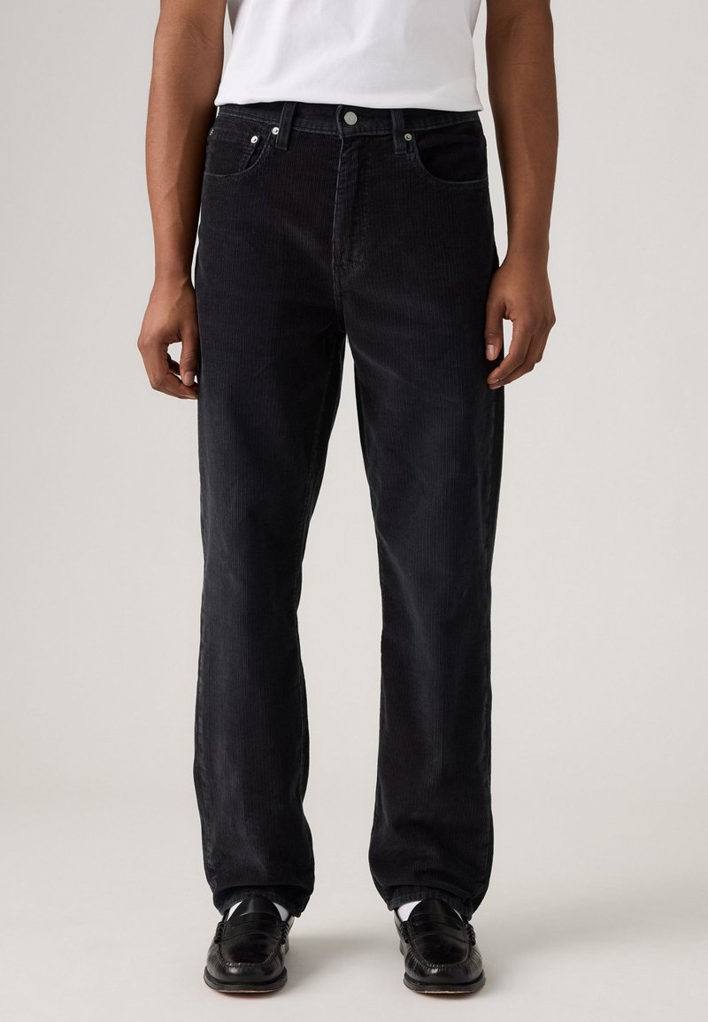 Zwarte corduroy broek met een rechte pasvorm, vijf zakken, metalen knoopsluiting en geribbelde textuur, gedragen met zwarte schoenen en een wit shirt.