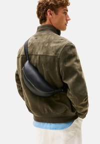 Borsa a tracolla in pelle nera indossata sopra una giacca in camoscio verde. La borsa presenta un design curvo con dettagli testurizzati e una singola tracolla regolabile.