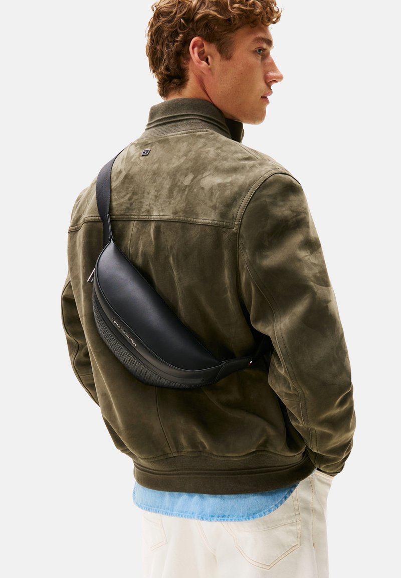 Borsa a tracolla in pelle nera indossata sopra una giacca in camoscio verde. La borsa presenta un design curvo con dettagli testurizzati e una singola tracolla regolabile.