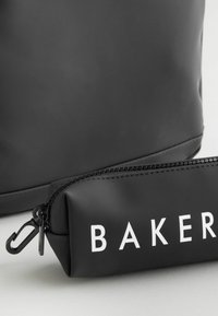 Crna torba s glatkom teksturom i zip-zatvorenim džepom označenim s "BAKER" u bijeloj boji. Džep ima srebrnu kopču i zaobljeni dizajn.