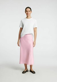 T-shirt blanc à manches courtes associé à une jupe rose mi-longue et des chaussures noires en maille à enfiler, le tout sur un fond clair uni.