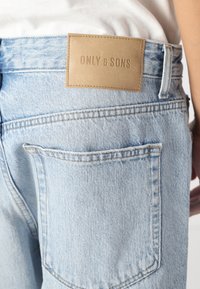 Jeans en denim bleu clair avec une finition texturée, dotés d'une patte en cuir marron avec le logo embossé "ONLY & SONS", et poches arrière standard.