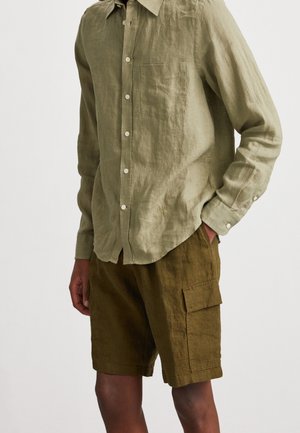 Mann trägt ein hellolivfarbenes langärmeliges Hemd mit Brusttasche und dunkelolivfarbene Cargoshorts mit seitlicher Klappentasche, Hände in den Taschen.