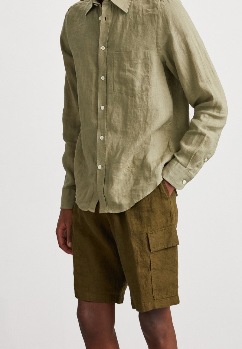 Homme portant une chemise boutonnée à manches longues couleur olive clair avec poche poitrine et un short cargo couleur olive foncé avec poche latérale à rabat, mains dans les poches.