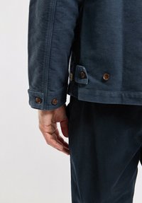Veste en velours côtelé bleu marine avec un col détaillé, des boutons aux poignets et des poches latérales, présentée avec une main posée sur le côté.