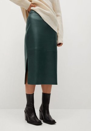 Femme portant une jupe midi en cuir vert foncé avec une fente latérale, un pull crème et des bottines à talons noires sur un fond uni.