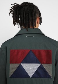 Grüne Jacke mit einem großen Aufnäher auf dem Rücken, der ein geometrisches Design in Rot, Marineblau, Grau und Weiß zeigt. Strukturierter Stoff und geschichtete Muster.