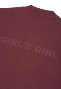 Ciemnoczerwony materiał z wypukłym ciemnym napisem "GIRL'S GIRL" umieszczonym na plecach, w pobliżu kołnierza ubrania.