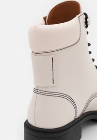 Bottine blanche en cuir épais avec surpiqûres noires contrastantes et bout carré. Présente une finition lisse et une étiquette texturée sur le côté.