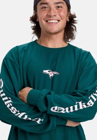 T-shirt à manches longues en coton vert avec le logo Quiksilver sur la poitrine et un texte blanc le long des manches, avec un col rond et une coupe décontractée.