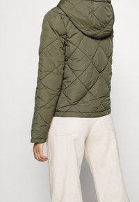 Pikowana kurtka oliwkowozielona z kapturem, ozdobiona wzorami diamentowymi i elastycznymi mankietami. Do tego jasne spodnie z szerokimi nogawkami.