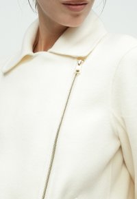 Hvid jakke i uldblanding med krave og gulddetalje i lynlåsen. Glat tekstur med skræddersyet pasform.