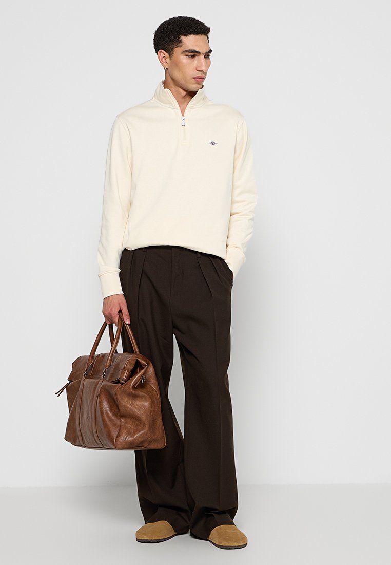 Cremefarbener Reißverschluss-Pullover, dunkle Hosen und braune Ledertasche, mit strukturierten Materialien und lockerer Passform. Braune Wildlederschuhe.