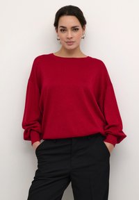 Crvena pletena pulover s okruglim izrezom i širokim rukavima, nosi se preko crnih hlača. Materijal ima blagi sjaj.