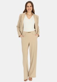 Beżowy żakiet dopasowany noszony na białej koszuli z dekoltem w serek, w połączeniu z szerokimi beżowymi spodniami z paskiem w złotym kolorze. Gładki materiał, zorganizowany fason.