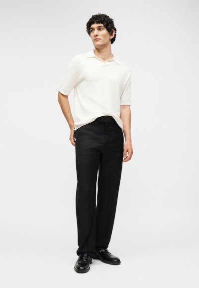 Άνδρας στέκεται φορώντας λευκό πόλο μπλουζάκι με κοντά μανίκια, μαύρο παντελόνι καλοραμμένο και μαύρα επίσημα παπούτσια σε απλό φόντο.