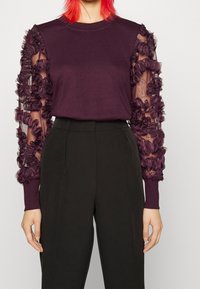 Donna che indossa un top viola con maniche trasparenti ricamate in 3D a fiori e pantaloni neri a vita alta, in piedi di fronte a uno sfondo semplice.