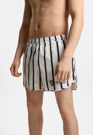 Męski tułów ubrany w białe kąpielówki w czarne pionowe paski z elastycznym pasem, stojący na jednolitym tle.