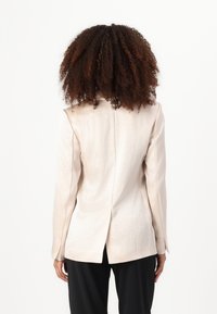 Svijetlo bež blazer od teksturirane tkanine s otvorenim reverima i stražnjim prorezom. Upareno s crnim hlačama koje ističu elegantan, krojeni dizajn.