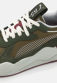 Gros plan d'une basket Puma vert olive et marron avec des lacets blancs, un design rayé sur le côté et une semelle blanche rembourrée avec une semelle bordeaux.