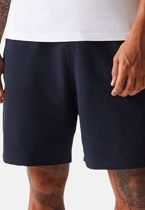 Mann trägt schlichte dunkelblaue Shorts und ein weißes Oberteil, mit sichtbaren Tattoos am linken Arm und an den Beinen.